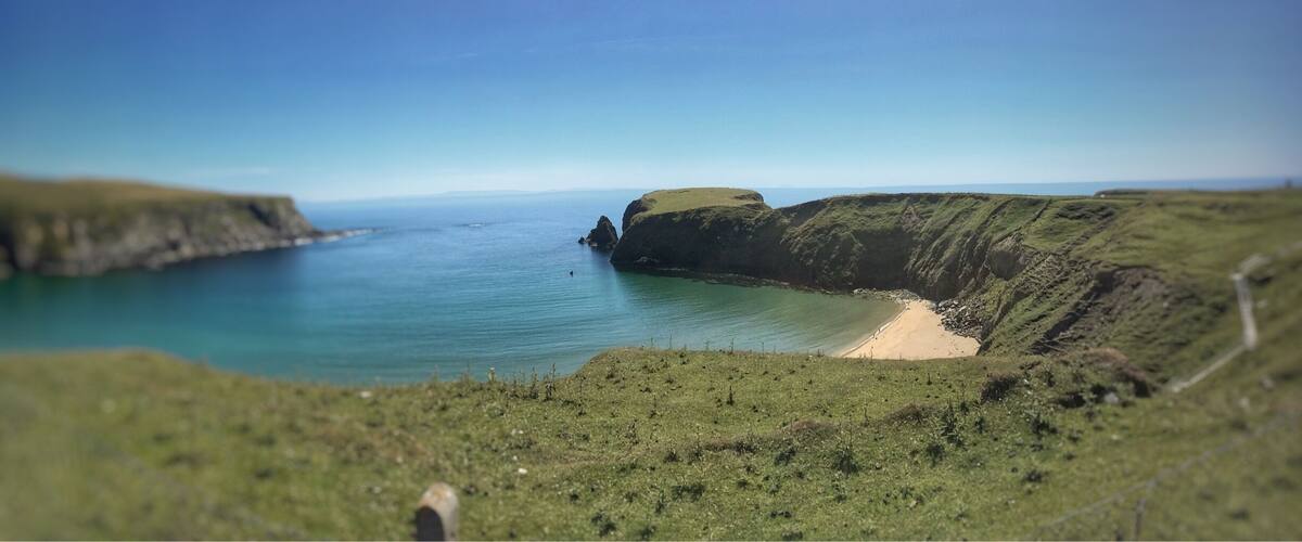 A coastal landscape with a sandy beach, grassy hills, and a calm sea.