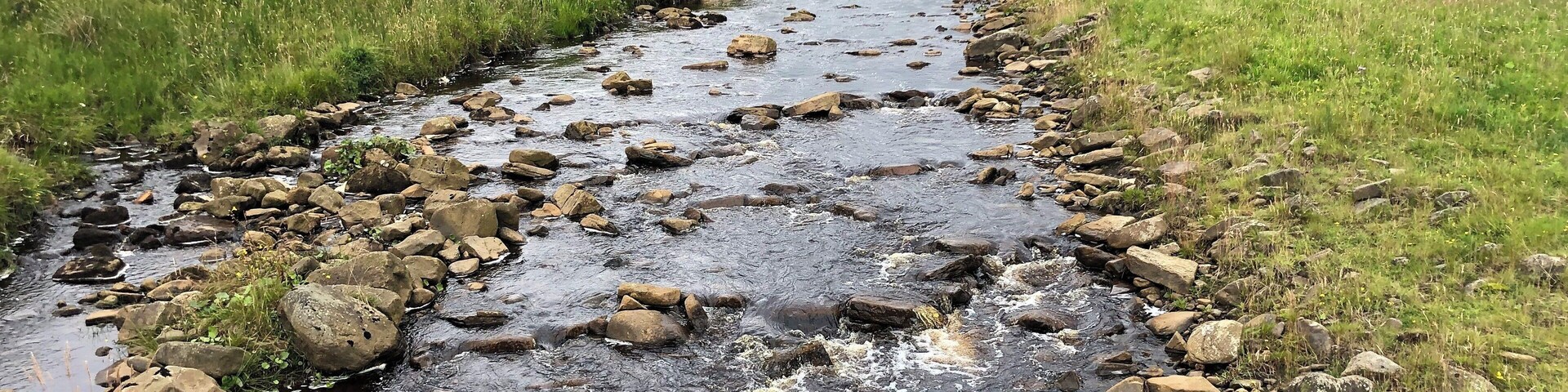 A rocky stream in a grassy valley with a stone bridge in the distance.