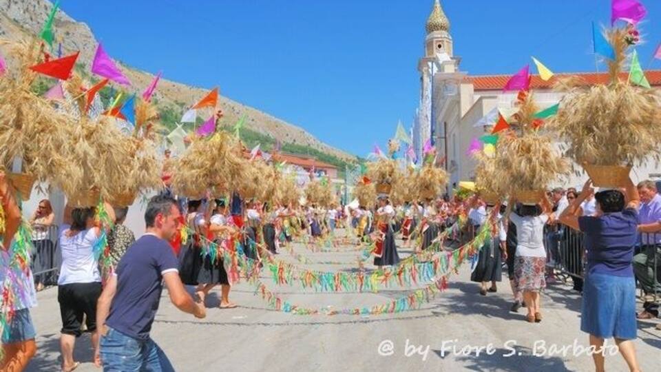 A festive procession with people carrying decorated baskets and colorful flags, leading towards a historic building with a clock tower.