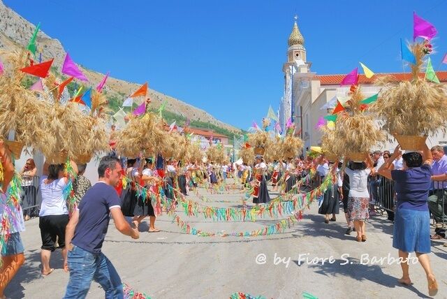 A festive procession with people carrying decorated baskets and colorful flags, leading towards a historic building with a clock tower.