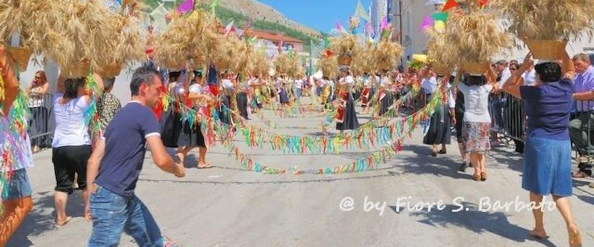 A festive procession with people carrying decorated baskets and colorful flags, leading towards a historic building with a clock tower.