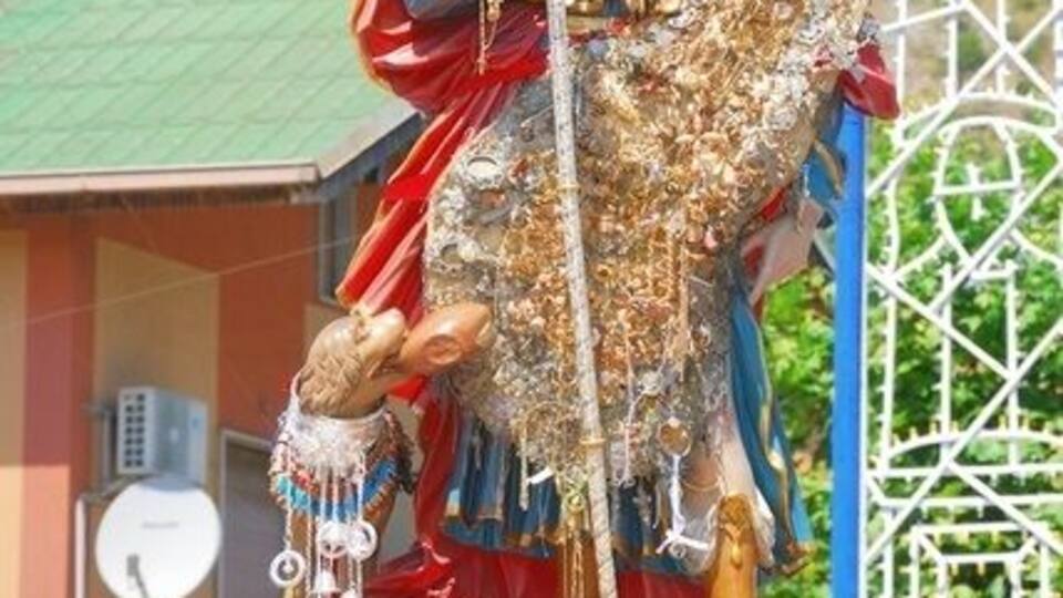 A statue of a religious figure with a halo, holding a staff and a book, adorned with a crown and jewels, set against a mountainous backdrop.