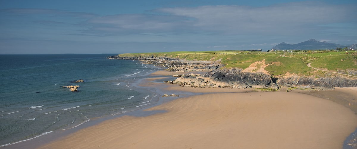 A coastal landscape with a sandy beach, rocky outcrops, and a distant mountain range.
