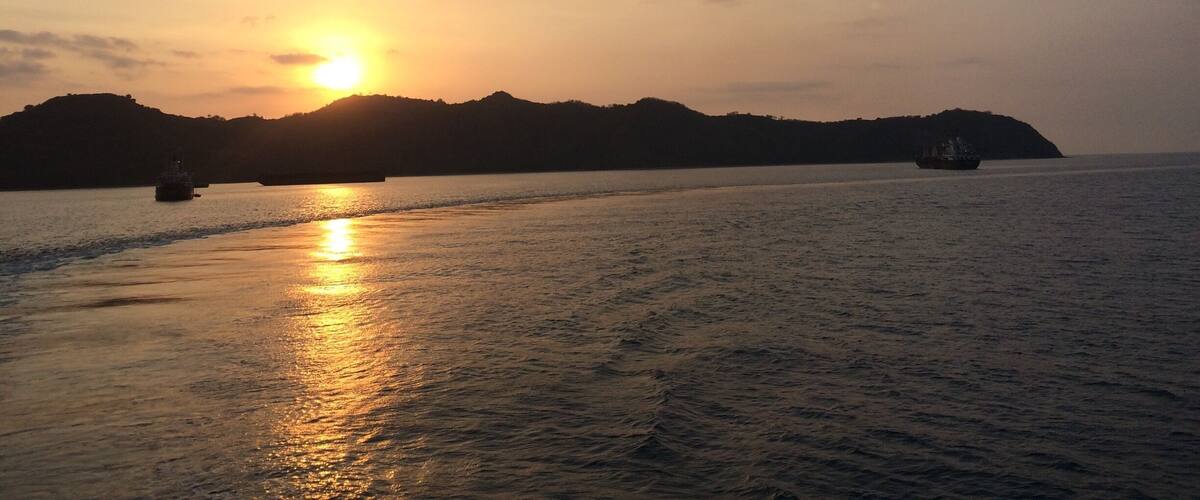 A sunset over a calm sea with boats and distant hills.