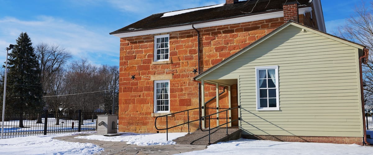 A brick house with a small attached shed, a snow-covered yard, and a clear blue sky.