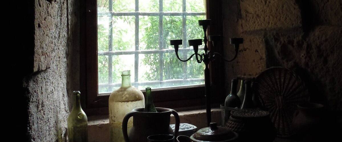 A dimly lit room with a large arched window, a candelabra, and various antique kitchen items.