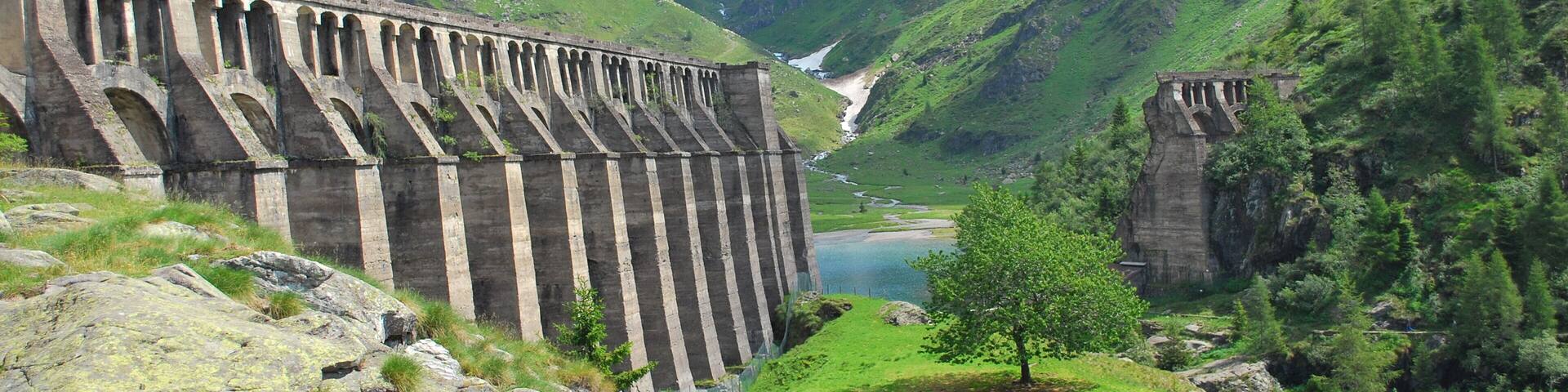 A dam with multiple arches in a mountainous valley with a lake and snow-capped peaks in the background.