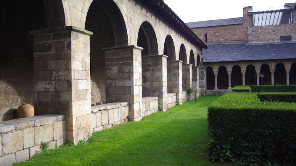 A stone building with arched walkways and a well-maintained hedge.