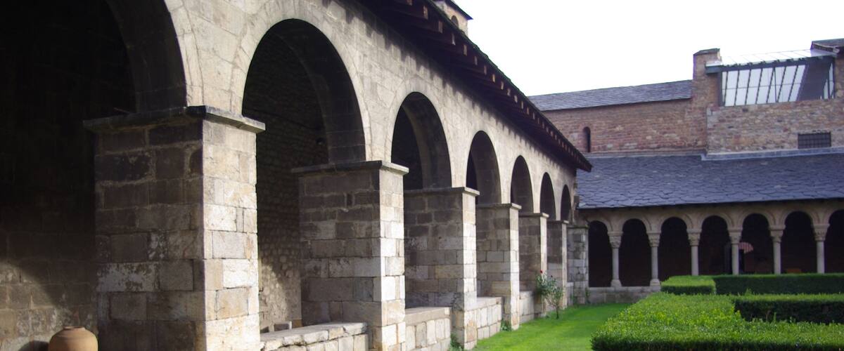A stone building with arched walkways and a well-maintained hedge.