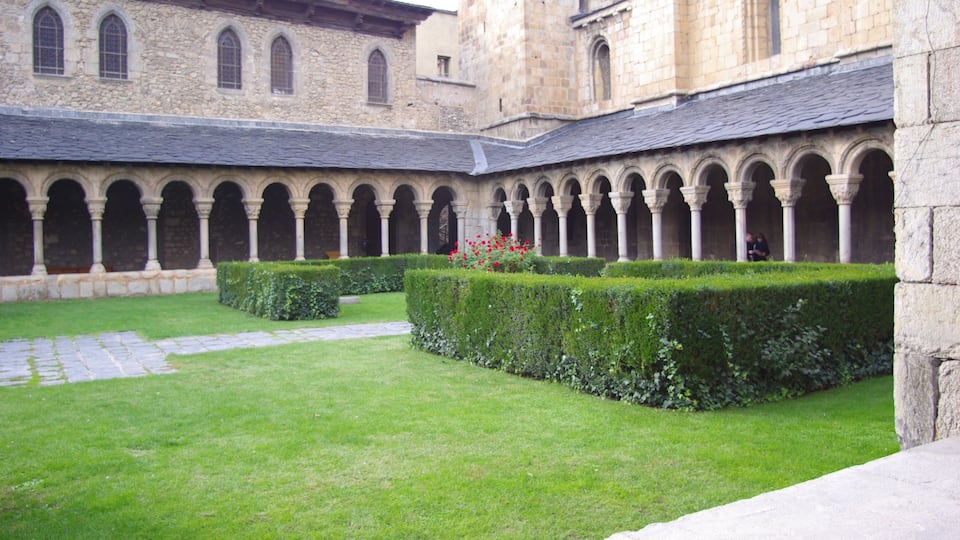 A courtyard with a stone building featuring arched walkways and a well-maintained hedge.