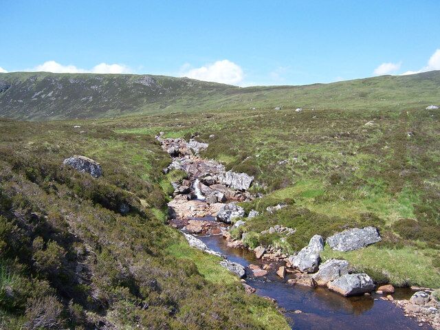 Un arroyo serpentea por un paisaje cubierto de hierba, con afloramientos rocosos y colinas al fondo.