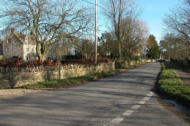 A quiet rural road lined with trees and a stone wall, leading to a stone house.
