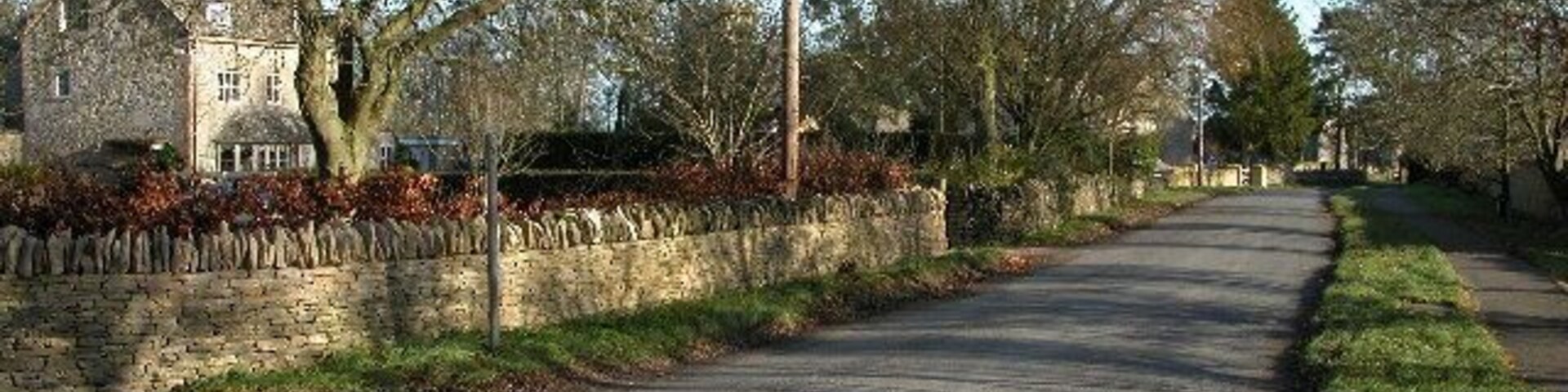 A quiet rural road lined with trees and a stone wall, leading to a stone house.