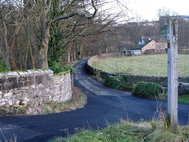 A narrow rural road winding through a stone wall and a grassy field with a building in the distance.