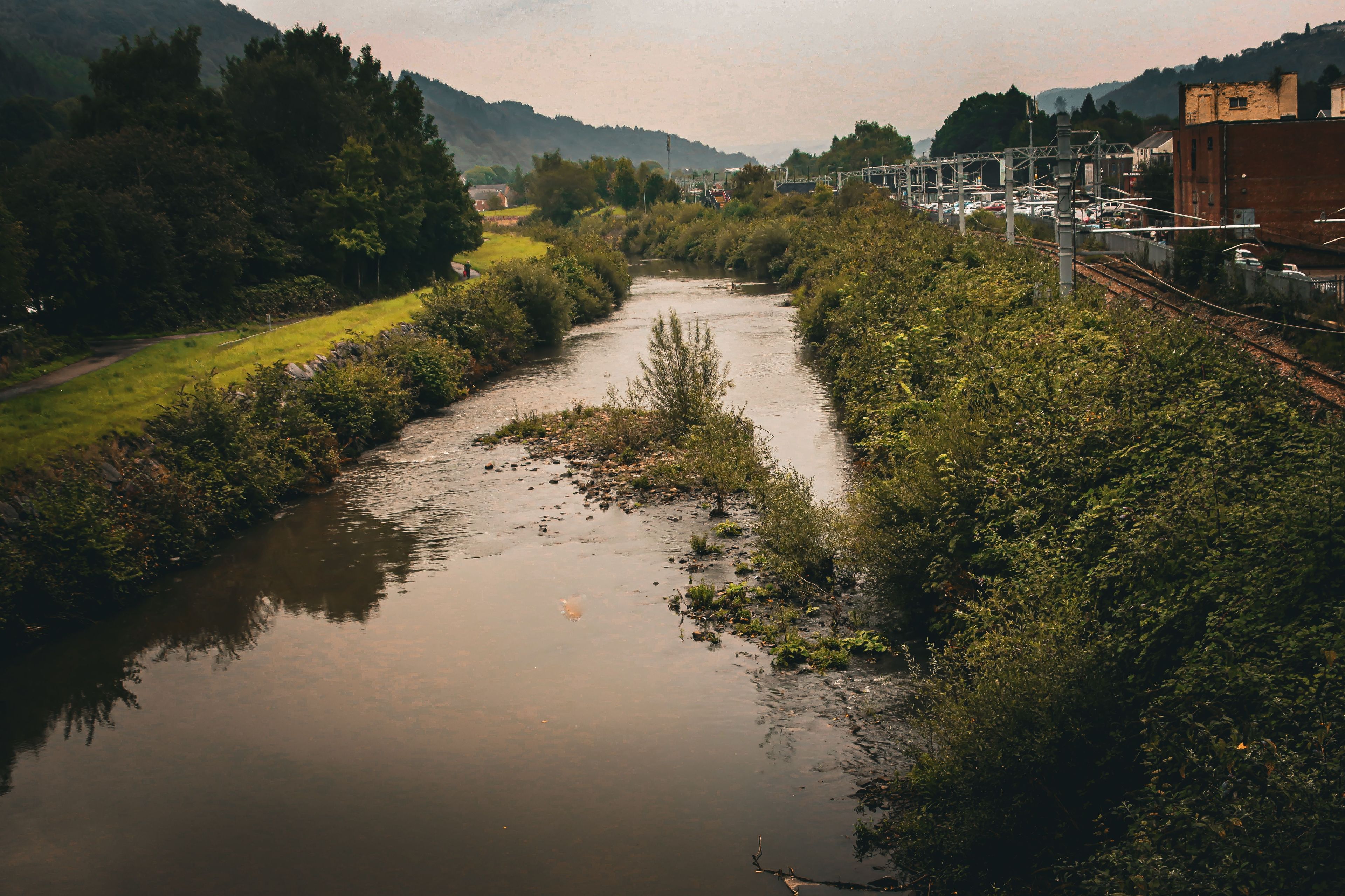 A river flowing through a rural area with greenery on both sides, a bridge, and a railway track.