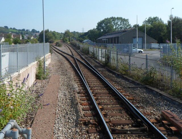 Trilhos de trem com uma cerca à esquerda, um edifício à direita e casas ao fundo.