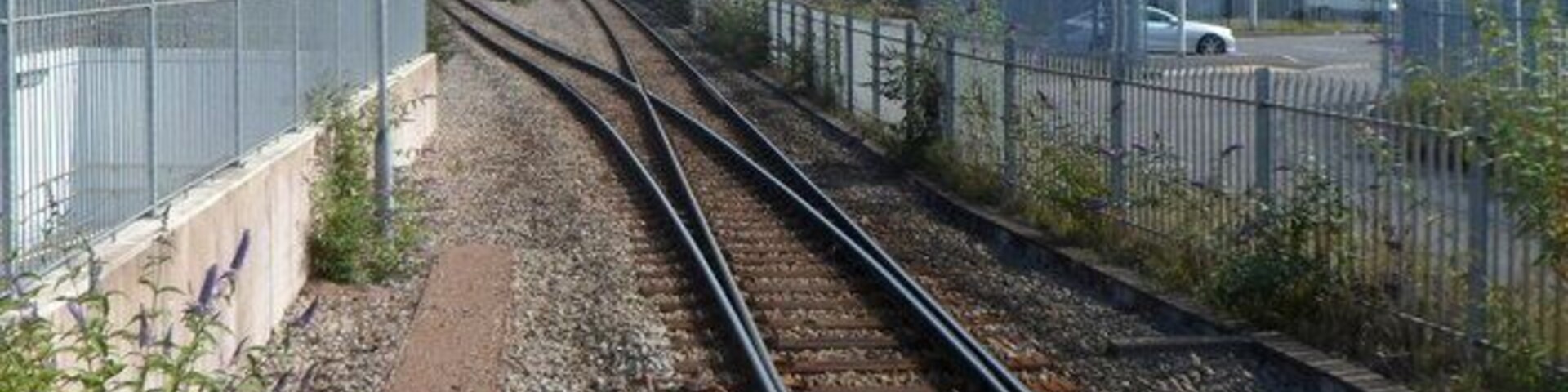 Trilhos de trem com uma cerca à esquerda, um edifício à direita e casas ao fundo.