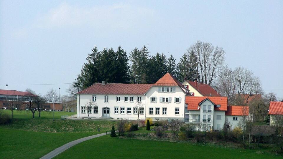 A large white building with a red roof, surrounded by green fields and trees.