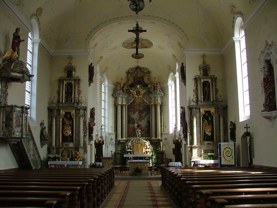 Interior d'una església amb detalls ornamentals, pews de fusta, un altar central i estàtues religioses.