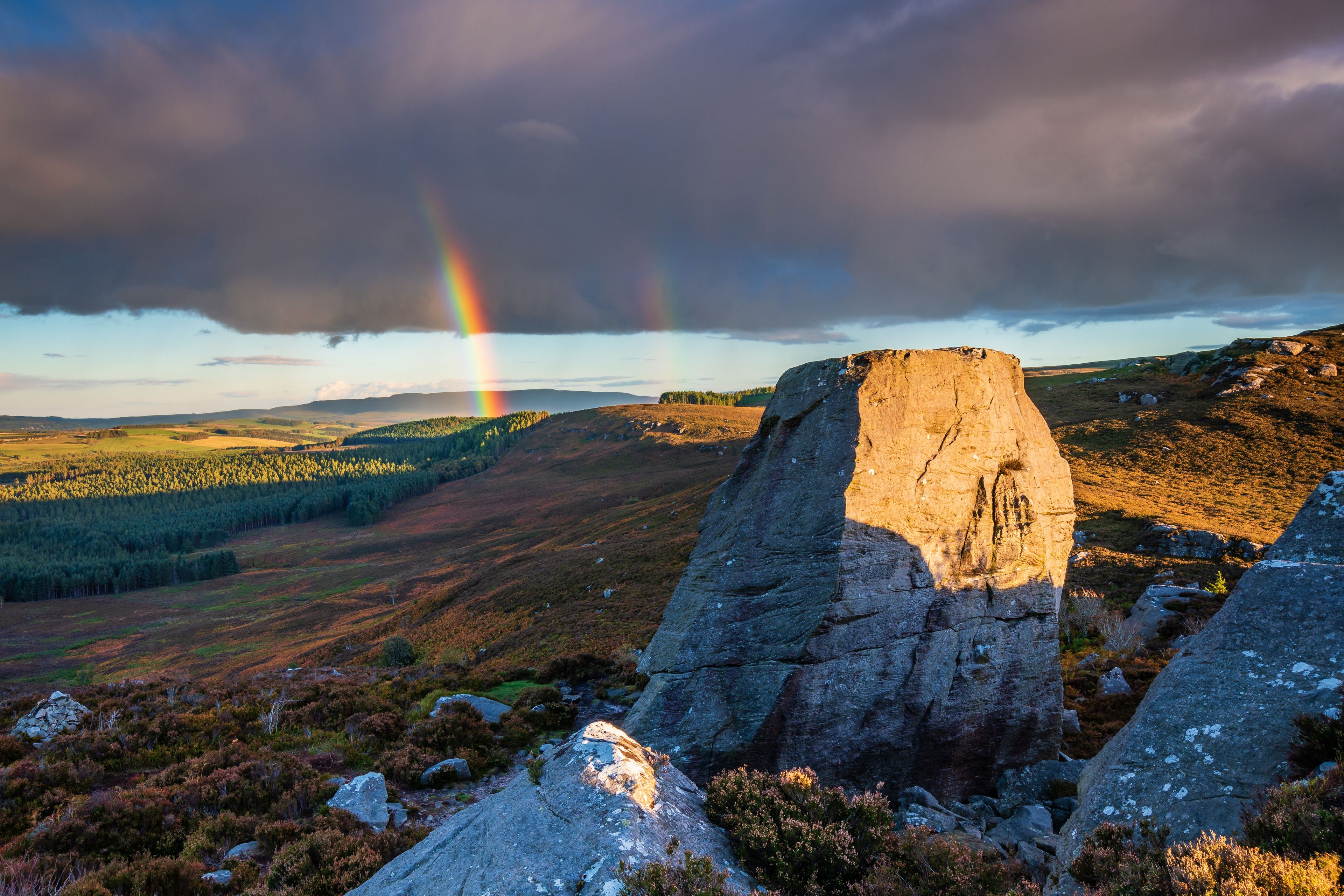 A rocky landscape with a prominent rock formation in the foreground, a forested valley in the midground, and a rainbow in the background.