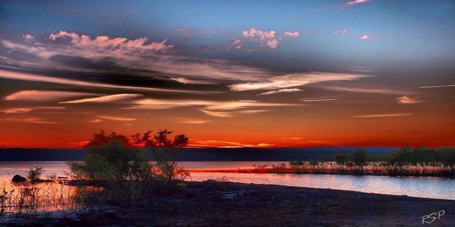 A sunset over a calm lake with silhouettes of trees and a clear sky.