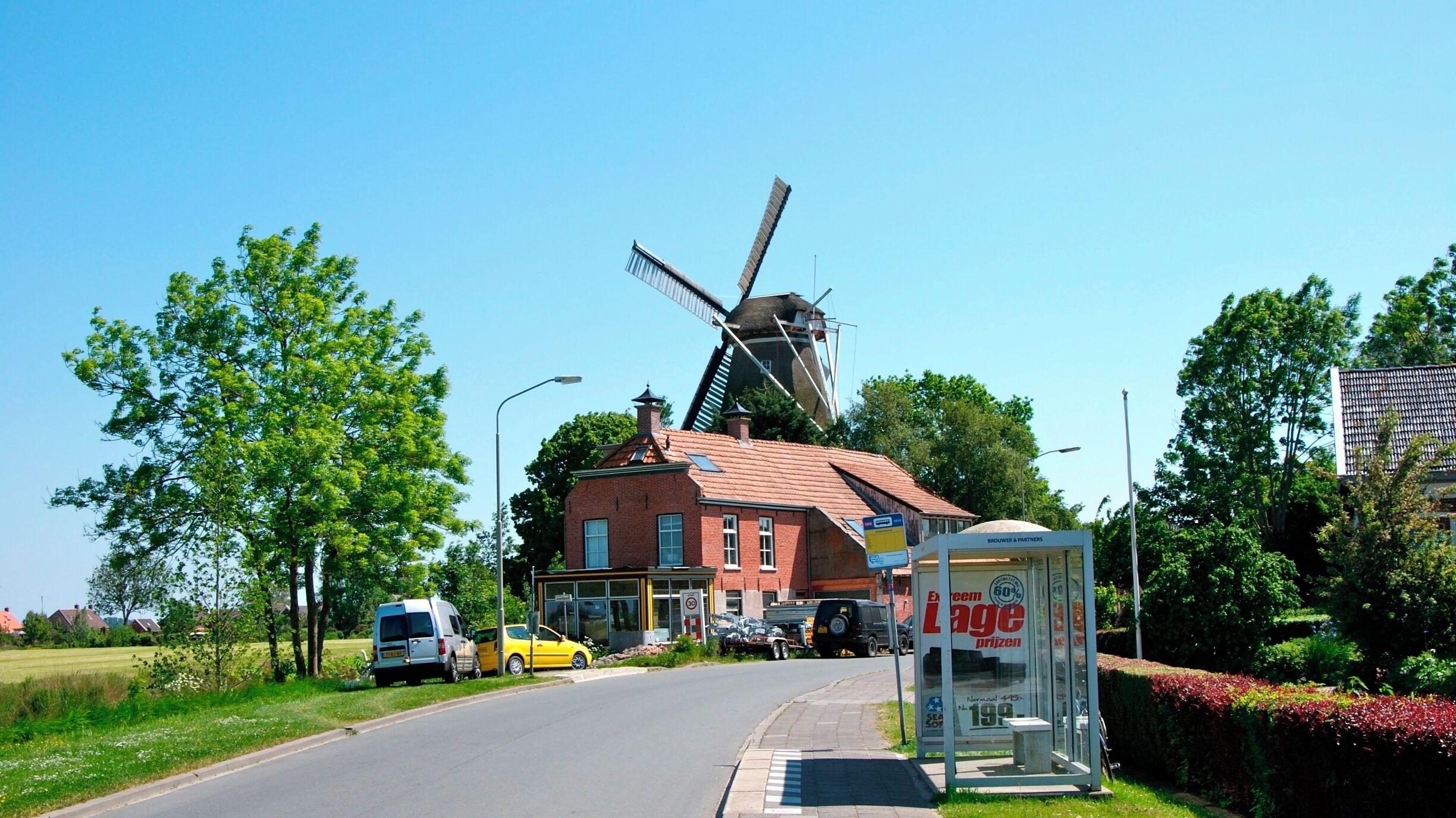A rural road with a windmill, a red brick building, and a bus stop.