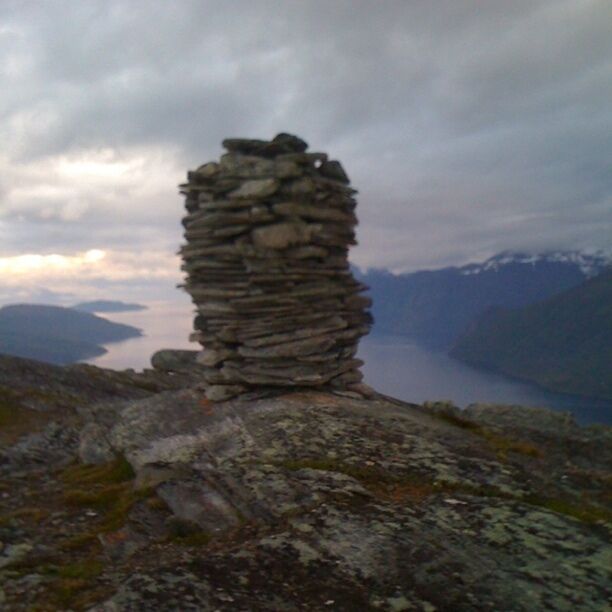 A stone cairn on a rocky outcrop with a lake and mountains in the background.