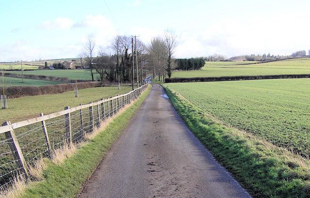A rural road flanked by fields and a fence.