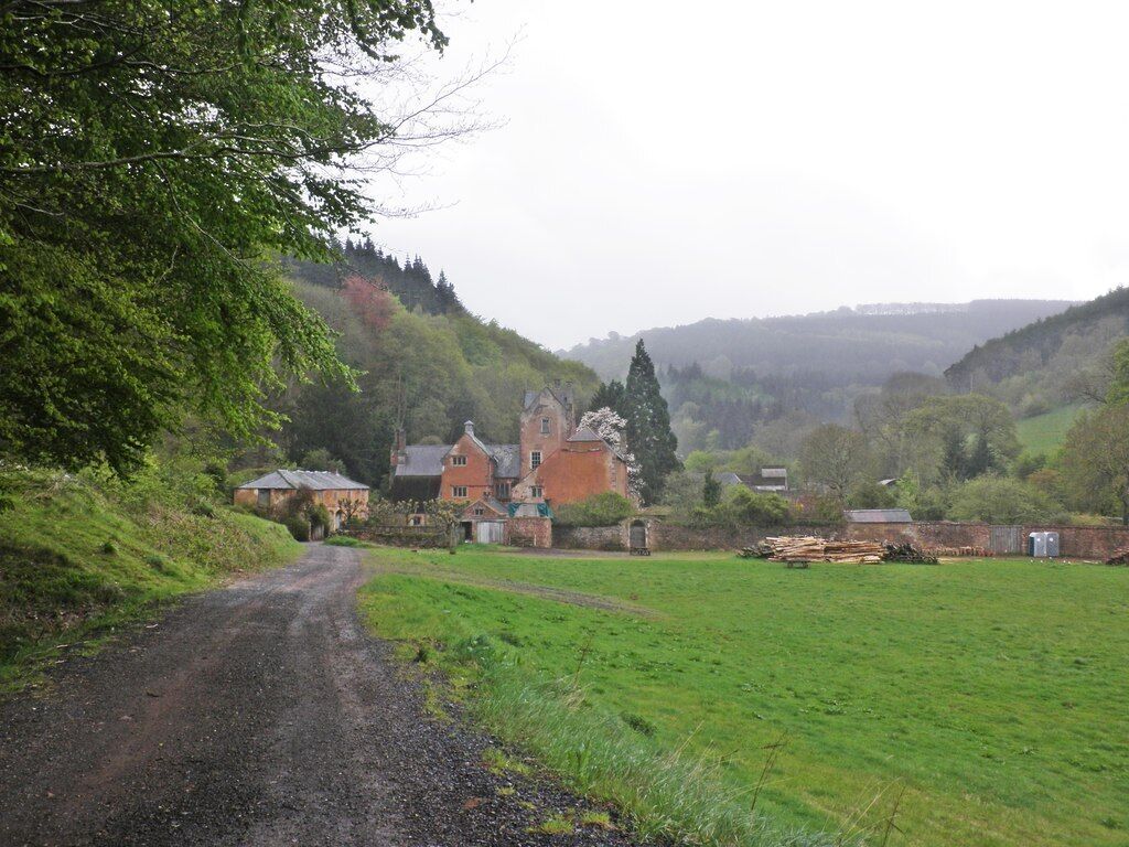A rural landscape with a gravel road leading to a large estate with multiple buildings, surrounded by lush greenery and hills in the background.