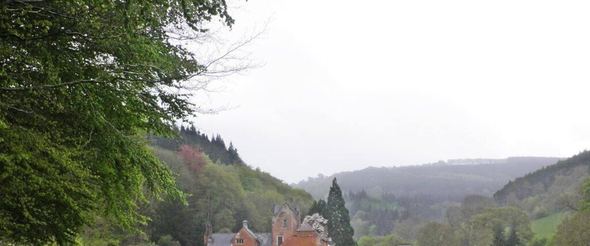 A rural landscape with a gravel road leading to a large estate with multiple buildings, surrounded by lush greenery and hills in the background.