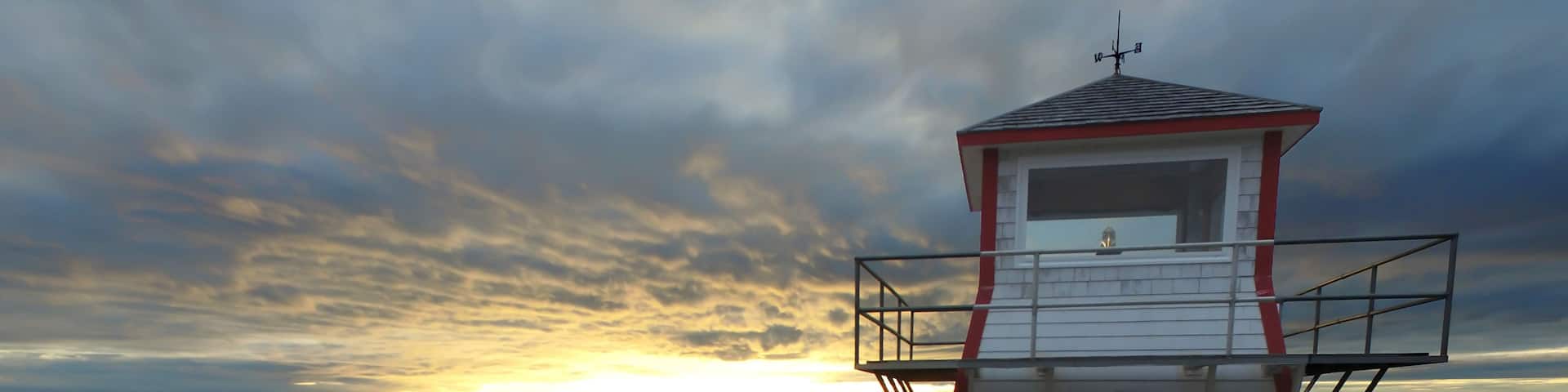 A white lighthouse with a red trim, situated on a rocky shoreline with a view of the sunset over the ocean.