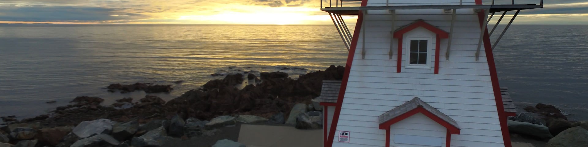 Un faro blanco con detalles rojos, situado en una costa rocosa con vista al atardecer sobre el océano.