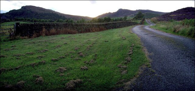A rural road curving through a green field with stone walls and hills in the background.