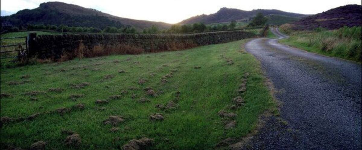 A rural road curving through a green field with stone walls and hills in the background.