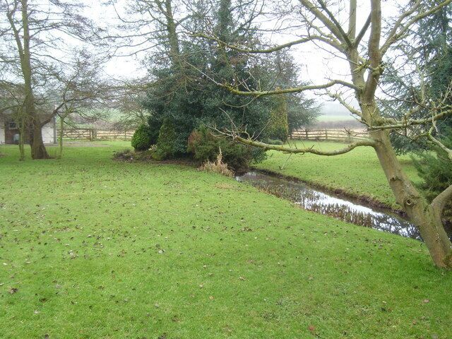 A grassy field with a small stream, trees, and a distant building.