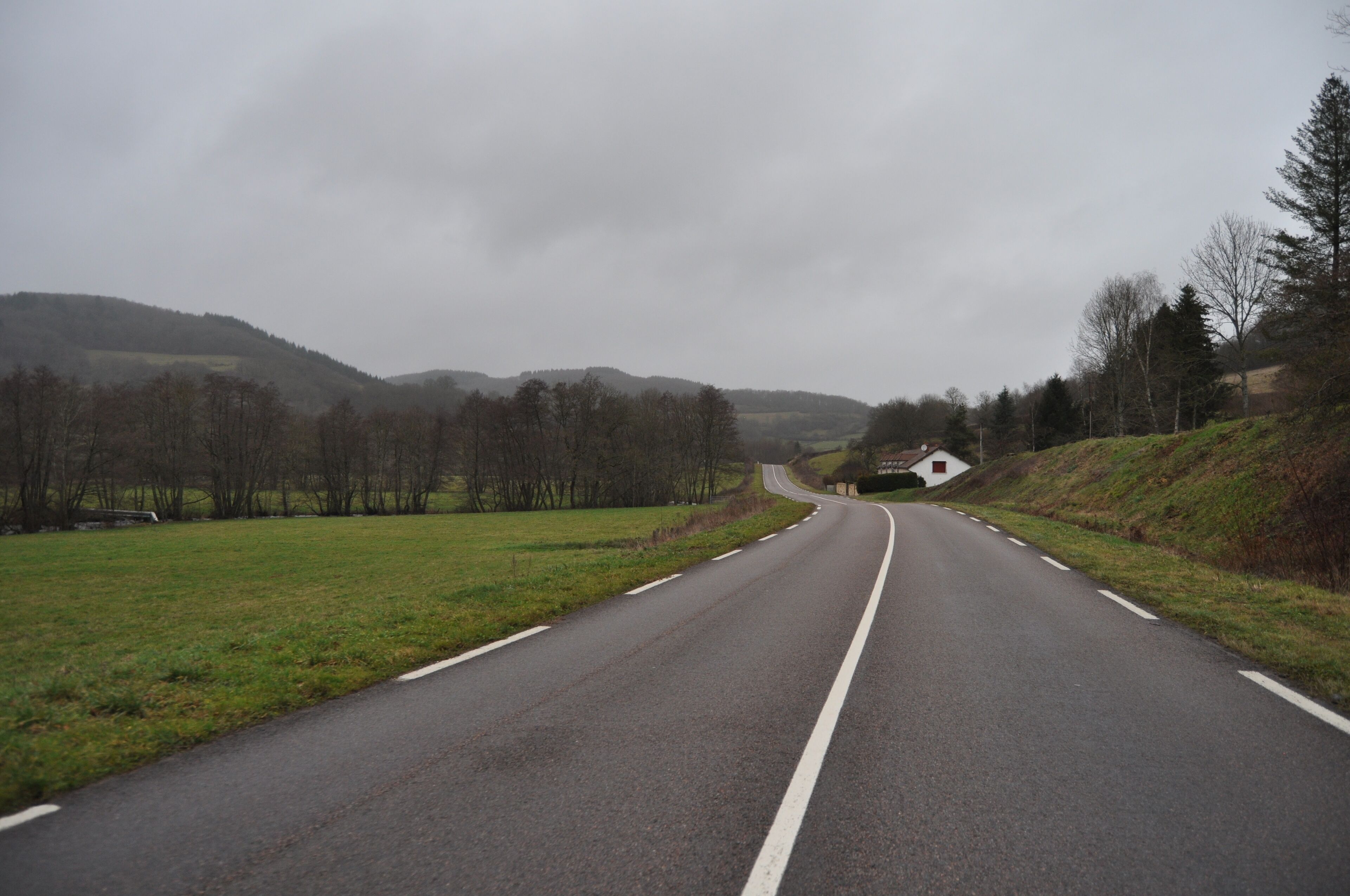 A rural road curving through a hilly landscape with sparse trees and a small house on the right.