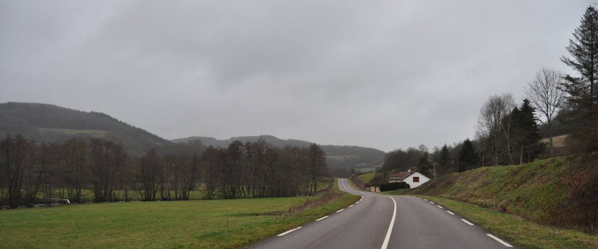 A rural road curving through a hilly landscape with sparse trees and a small house on the right.