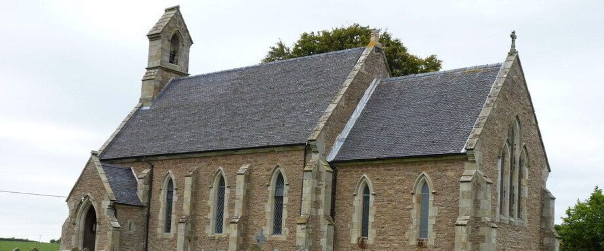 A stone church with a steep roof and multiple windows, surrounded by a cemetery with headstones.