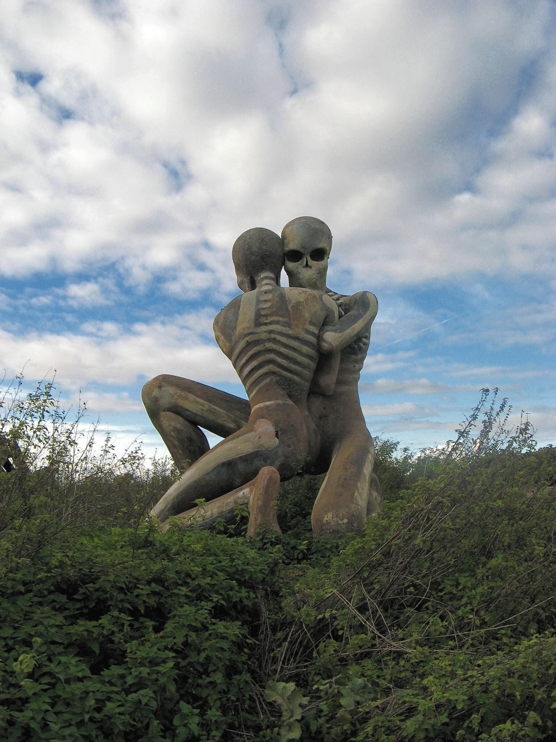 A large skeleton sculpture of a man and woman embracing, set against a cloudy sky and surrounded by greenery.