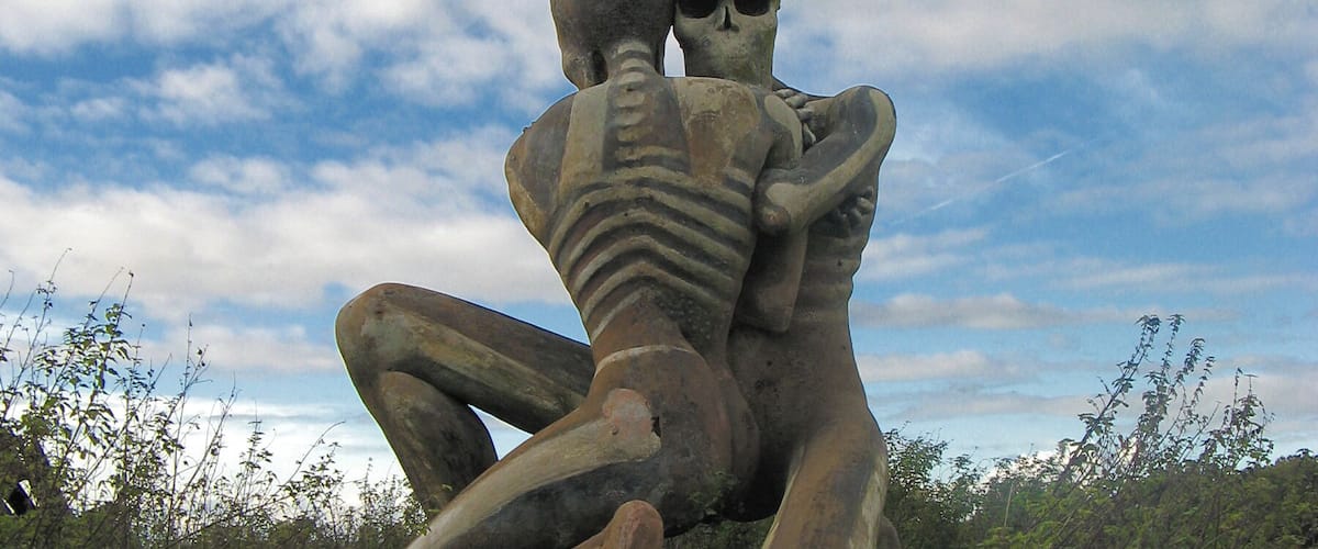 A large skeleton sculpture of a man and woman embracing, set against a cloudy sky and surrounded by greenery.