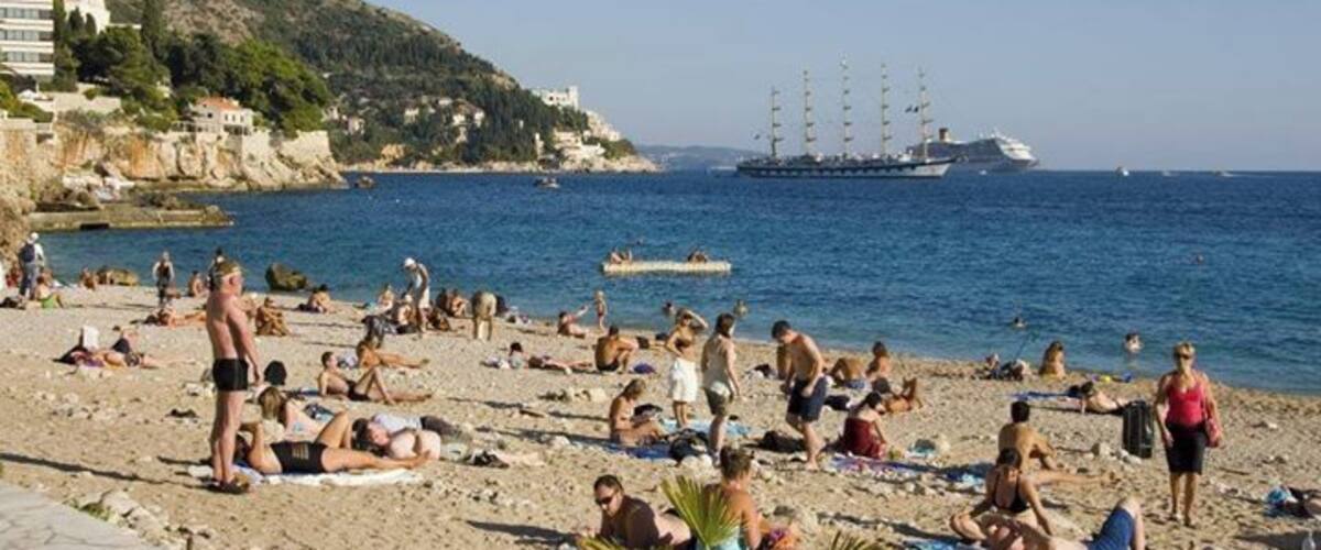 A crowded beach with people sunbathing and swimming, a large cruise ship in the background, and a hilly landscape.