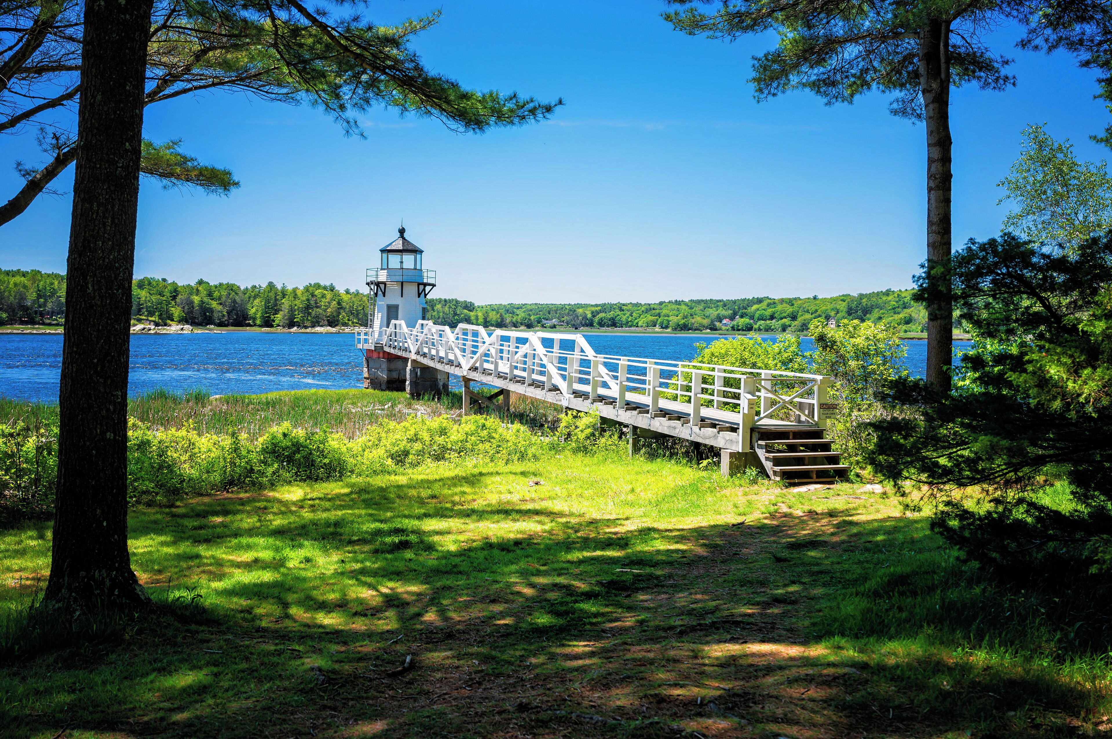 A wooden bridge with a white railing leading to a lighthouse by a lake surrounded by trees.