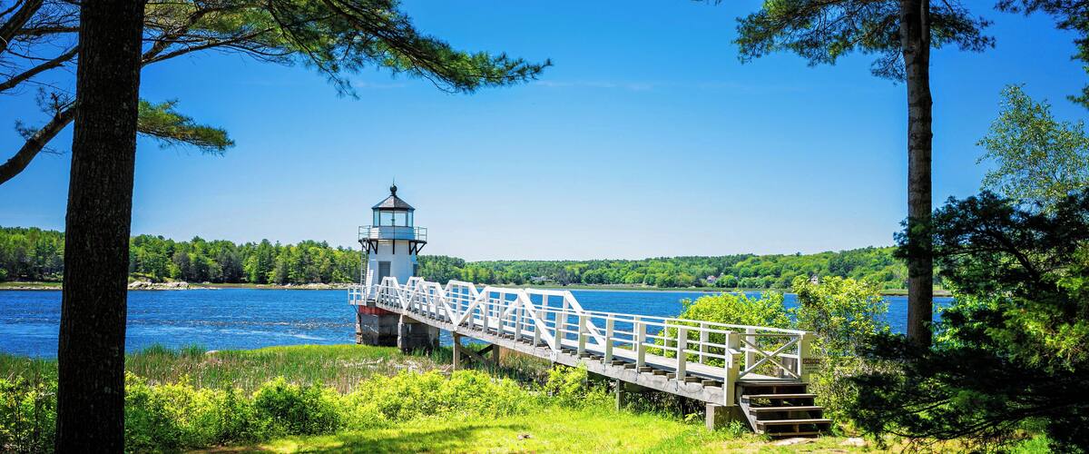 A wooden bridge with a white railing leading to a lighthouse by a lake surrounded by trees.