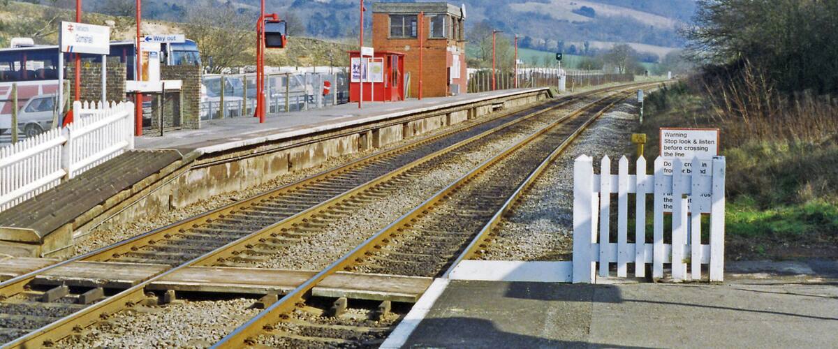 A railway station platform with tracks, a signal box, and a white picket fence.