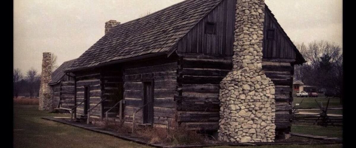 A rustic log cabin with a stone chimney, surrounded by a grassy field and a stone wall.