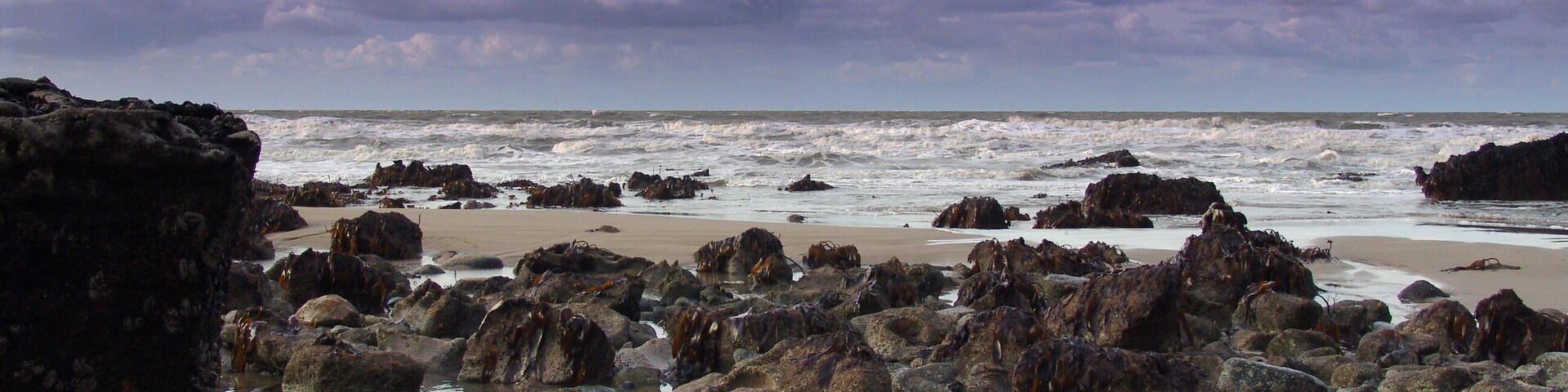 A rocky shoreline with waves crashing against the rocks and a beach in the background.