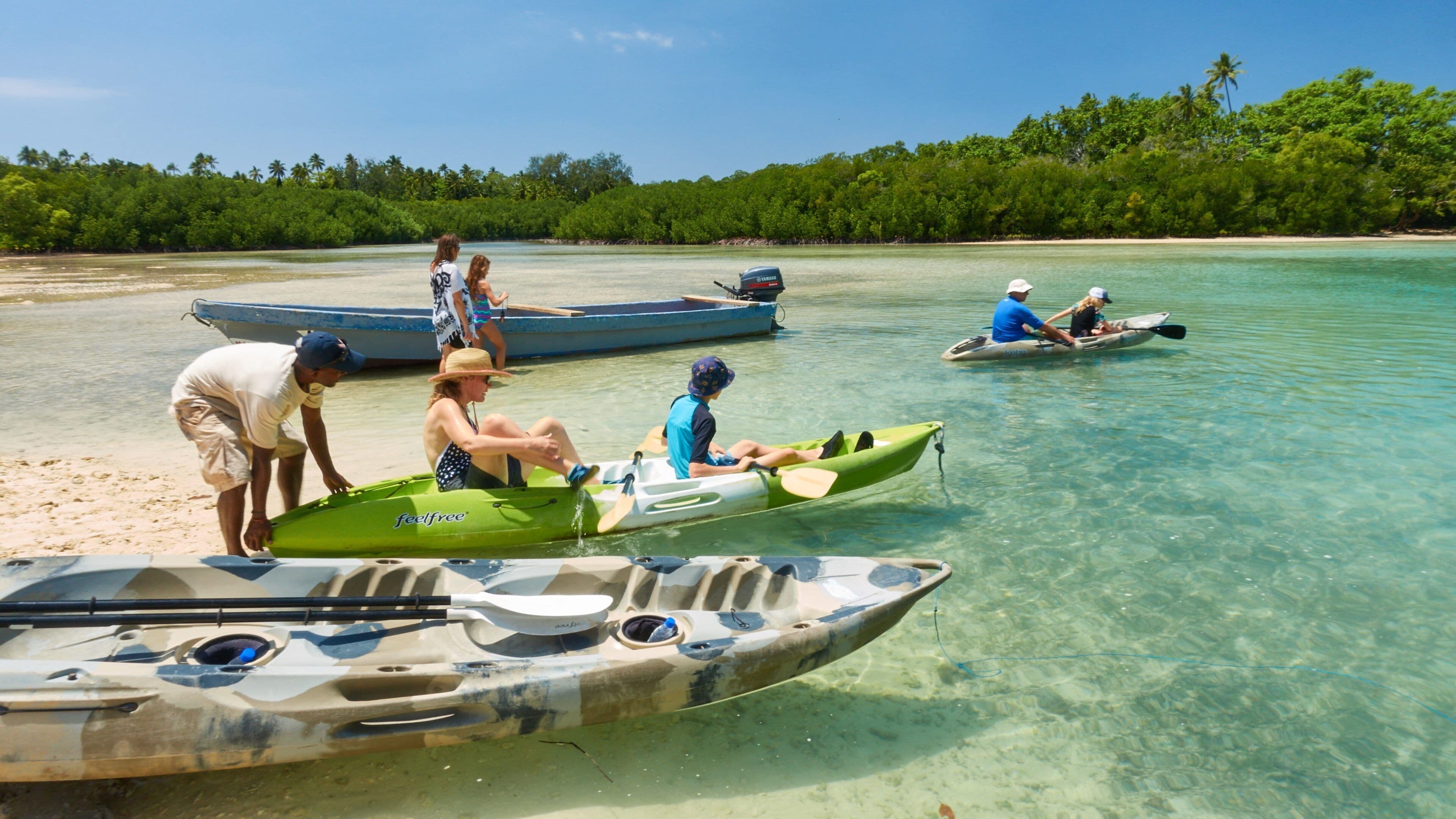 Un grupo de personas preparando kayaks en una playa con agua turquesa cristalina y un fondo arbolado.