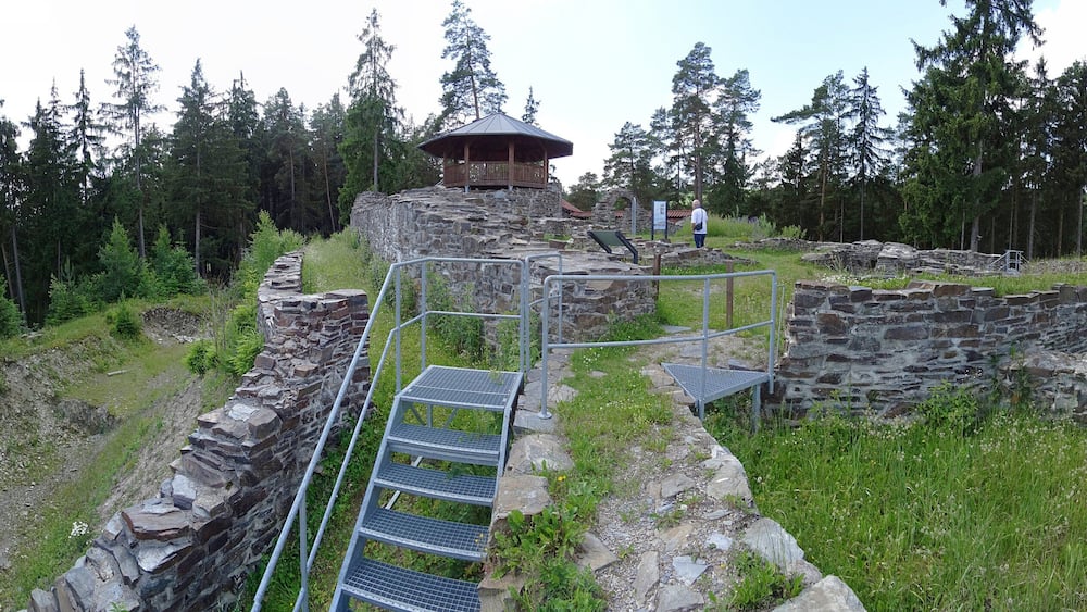 A stone ruin with a wooden viewing platform and metal stairs leading up to it, surrounded by a forest.