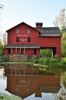 Daytime photo of the red barn mill at Bonneyville Mills in Bristol, Indiana with a calm pond and reflection in the foreground