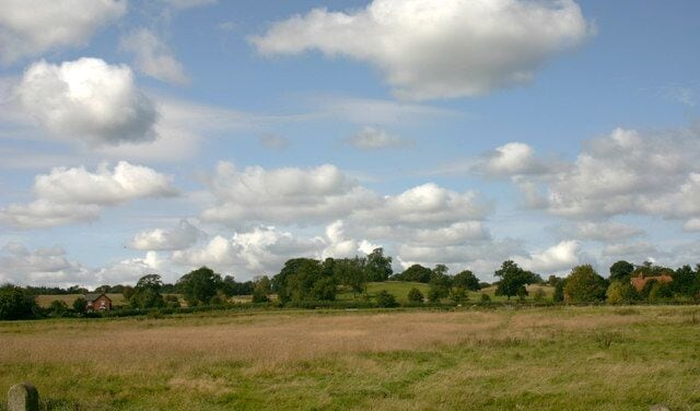 A rural landscape with a field, scattered trees, and a partly cloudy sky.